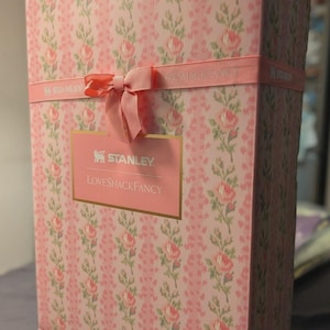 Puede incluir: Una caja de regalo rectangular con un estampado floral rosa y una cinta rosa atada con un lazo. La caja presenta los logotipos "STANLEY" y "LOVESHACKFANCY" en letras doradas sobre una etiqueta rosa.