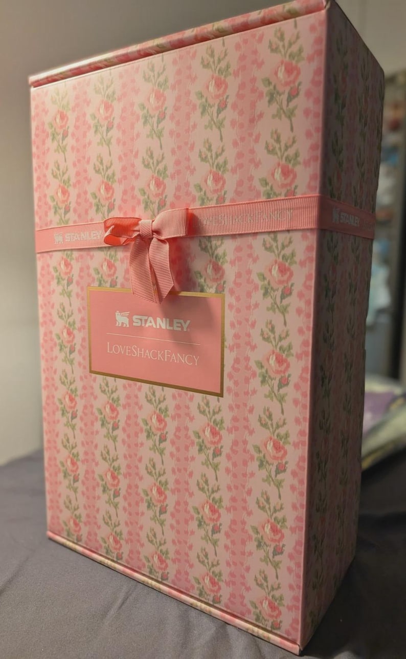 Puede incluir: Una caja de regalo rectangular con un estampado floral rosa y una cinta rosa atada con un lazo. La caja presenta los logotipos "STANLEY" y "LOVESHACKFANCY" en letras doradas sobre una etiqueta rosa.
