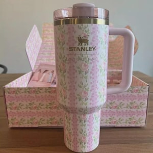Puede incluir: Un vaso Stanley con estampado floral, asa y tapa rosas. El vaso presenta un dise&ntilde;o floral rosa y blanco con el texto "STANLEY LOVESHACKFANCY". El vaso est&aacute; en una caja floral a juego.