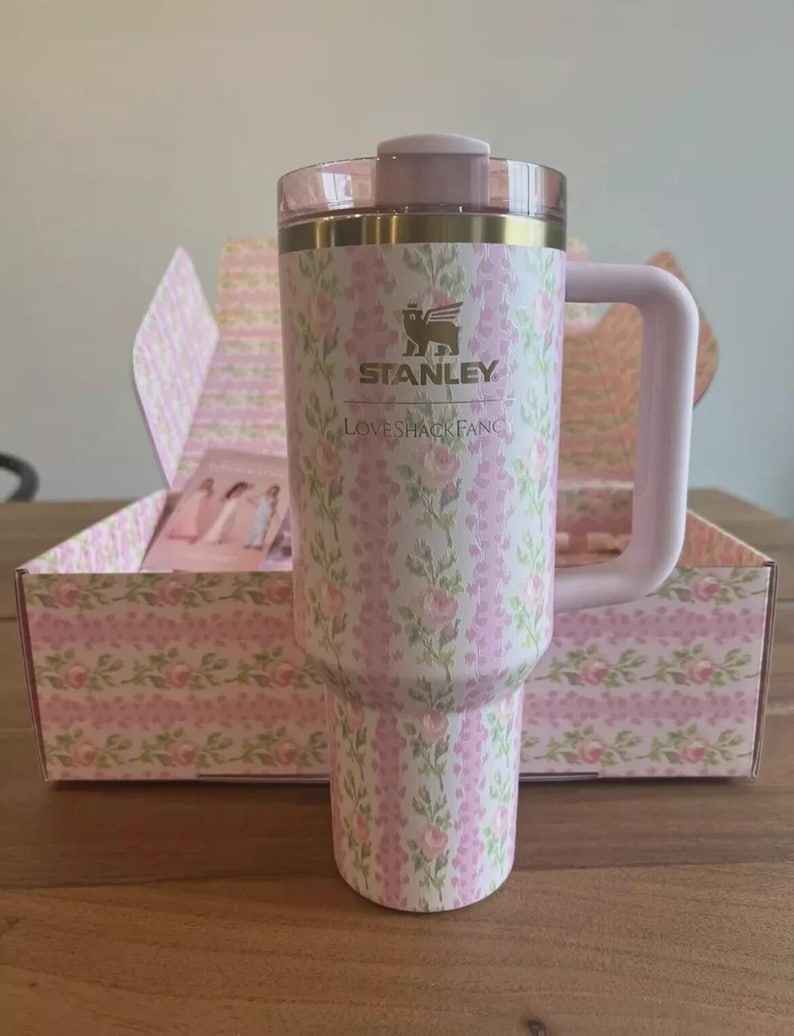 Puede incluir: Un vaso Stanley con estampado floral, asa y tapa rosas. El vaso presenta un dise&ntilde;o floral rosa y blanco con el texto "STANLEY LOVESHACKFANCY". El vaso est&aacute; en una caja floral a juego.