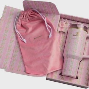Puede incluir: Un termo de viaje Stanley rosa con un estampado floral, asa y tapa, presentado en una caja de regalo a juego con una bolsa con cord&oacute;n rosa. La caja y la bolsa tambi&eacute;n presentan el dise&ntilde;o floral. El termo tiene un borde dorado.