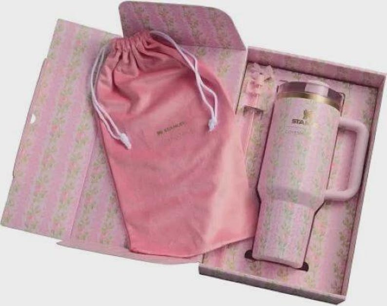 Puede incluir: Un termo de viaje Stanley rosa con un estampado floral, asa y tapa, presentado en una caja de regalo a juego con una bolsa con cord&oacute;n rosa. La caja y la bolsa tambi&eacute;n presentan el dise&ntilde;o floral. El termo tiene un borde dorado.