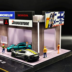 May include: A detailed miniature garage scene with a silver sports car and a teal sports car. The garage features signs for Honda, Bridgestone, and Michelin. Small figures are positioned around the cars.