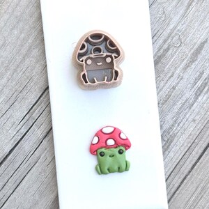 Puede incluir: Un cortador de galletas de plástico color canela con forma de rana con sombrero de hongo, colocado sobre una pequeña figura de arcilla terminada de una rana verde con un sombrero de hongo rojo con puntos blancos, montados en una tarjeta blanca.