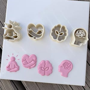 Puede incluir: Un juego de cuatro cortadores de galletas de plástico color crema y sus formas de fondant rosa correspondientes. Los diseños incluyen una persona en posición de meditación sentada, un corazón abrazado por dos brazos, un cerebro con un brote y un perfil de cabeza.