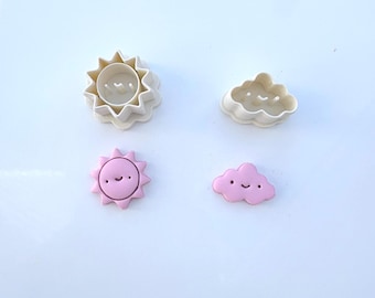 May include: Two off-white plastic cookie cutters shaped like a sun and a cloud, paired with two corresponding light pink fondant or clay shapes featuring embossed smiling faces with two eyes and a curved mouth on each piece.