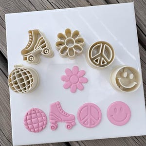 Può includere: Una collezione di tagliabiscotti in plastica color crema e le corrispondenti forme in argilla rosa chiaro. Il set include disegni di un pattino a rotelle, un fiore, un simbolo della pace, una palla da discoteca e una faccina sorridente su piastrella.