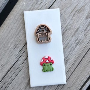 Peut inclure: Un emporte-pièce imprimé en 3D marron clair en forme de grenouille portant un chapeau de champignon, placé au-dessus d'une petite figurine en argile représentant une grenouille verte avec un chapeau de champignon rouge à pois blancs sur un carreau blanc.