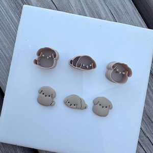 May include: A set of three light brown dog-shaped cookie cutters and three corresponding light brown dog-shaped fondant or dough cutouts. The cutters feature raised edges for shaping, while the cutouts display simple, embossed facial features.