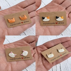 Op de afbeelding: Vier paar handgemaakte katvormige oorstekers op bruine kartonnen kaarten. De oorbellen hebben verschillende kleuren, waaronder oranje, zwart, wit en bruin. Elke kaart is voorzien van de tekst "Handmade with love."