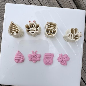 Puede incluir: Un juego de cuatro cortadores de galletas de plástico color crema y cuatro impresiones correspondientes en fondant o arcilla rosa claro. Las formas incluyen una colmena, una abeja, un tarro de miel y una abeja de perfil.