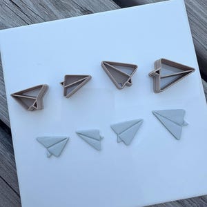 Peut inclure: Un ensemble de quatre emporte-pièces en plastique beige en forme d'avion en papier, disposés en ligne au-dessus de quatre formes d'avion en papier gris clair en pâte ou argile, sur une surface carrée blanche lisse.