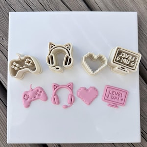 May include: A selection of gaming-themed cookie cutters in beige and pink. The shapes include a controller, headphones, a pixelated heart, and a computer monitor with the text "LEVEL UP!". The items are arranged on a white surface.