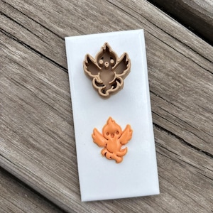 May include: A brown plastic cookie cutter shaped like a stylized bird with wings and a tail, paired with an orange clay or fondant cutout of the same bird design, both displayed on a rectangular white backing card.