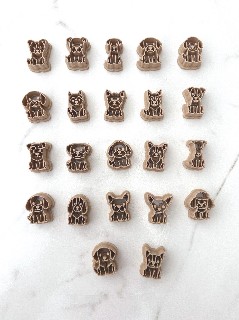 Twenty-two dog breed polymer clay cutters arranged in rows on a white marble surface showing a wide variety of sitting puppy designs.