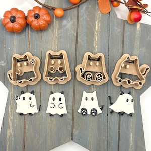 May include: Four tan plastic cookie cutters shaped like cats wearing ghost costumes, paired with four corresponding white and black cat ghost cookies. The set features various poses including waving, standing, and sitting, displayed on a gray wooden star.