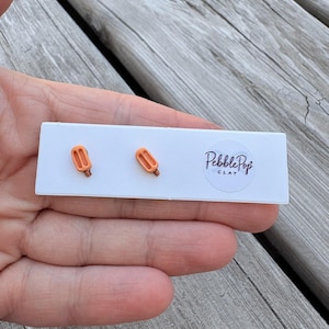Puede incluir: Un par de pequeños pendientes de botón con forma de polo naranja sujetos a una tarjeta de presentación rectangular blanca. La tarjeta presenta el texto Pebble Pop CLAY impreso en tinta negra sobre una pegatina circular blanca.