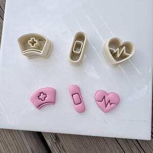 May include: Three beige plastic cookie cutters shaped like a nurse cap, a bandage, and a heart with an EKG line, alongside three corresponding pink fondant or clay cutouts of the same shapes, all arranged on a smooth white square surface.