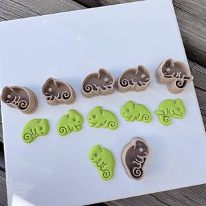 May include: A collection of chameleon-shaped cookie cutters in light brown plastic and corresponding light green chameleon-shaped fondant or dough cutouts. The set includes various sizes and poses of the reptile, featuring detailed embossed eyes and tails.