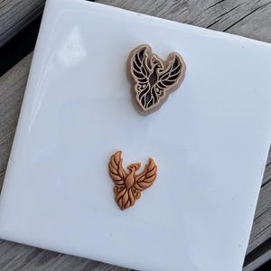 Può includere: Un tagliabiscotti a forma di fenice color beige e un timbro per biscotti più piccolo abbinato color rame. Entrambi gli oggetti presentano dettagli intricati e stilizzati sulle ali e sulle piume della coda, con un design simmetrico.