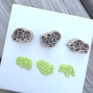 May include: Three light brown plastic turtle-shaped cookie cutters are arranged in a row above three corresponding light green turtle-shaped cutouts. Each turtle features a simple, rounded design with a small head, shell, and limbs.