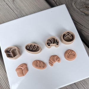 May include: A set of eight light brown plastic cookie cutters and matching stamps in various bread and pastry shapes, including a loaf of bread, an oval roll, a croissant, and a round bun, arranged on a white surface for baking and crafting.