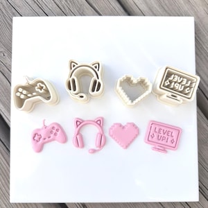 May include: A set of four beige plastic cookie cutters and four corresponding pink fondant shapes. The designs include a game controller, cat-ear headphones, a pixelated heart, and a computer monitor with the text LEVEL UP! and two diamond icons.