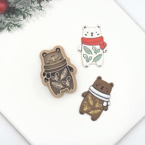May include: Three bear-shaped pins or ornaments featuring winter themes. One is a brown bear with a tan scarf and gold foliage, one is a white bear with a red scarf and green foliage, and one is a dark brown bear with a white scarf and gold foliage.