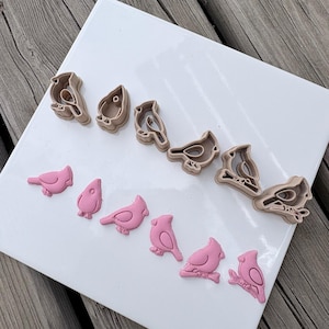 Puede incluir: Un conjunto de seis cortadores de galletas de plástico de color marrón claro con forma de pájaro dispuestos en fila, acompañados de seis recortes de fondant o arcilla rosa correspondientes en una fila paralela sobre una baldosa blanca.
