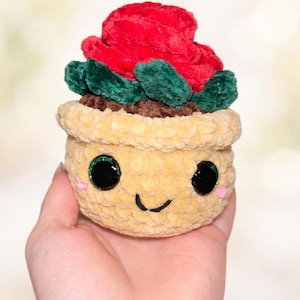 Peut inclure: Une plante succulente en peluche faite à la main dans un pot jaune avec un visage souriant. La plante succulente a une fleur rouge et des feuilles vertes. Le pot a des yeux noirs avec des étincelles vertes et des joues roses.