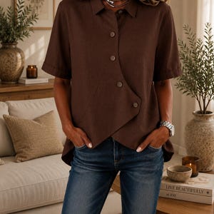 May include: A dark brown short-sleeved shirt with a collar and an asymmetrical button-down front closure. The shirt features a layered hemline and is paired with blue denim jeans. The fabric has a visible texture, and the buttons are round.