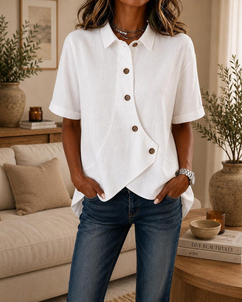 May include: A white short-sleeved button-down shirt featuring a unique asymmetrical front placket with brown buttons. The shirt has a classic collar, cuffed sleeves, and a hemline that angles downward, paired with dark blue denim jeans.