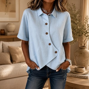 May include: A light blue short-sleeved shirt with a collar and an asymmetrical button-down front featuring five brown buttons. The shirt has a straight hemline and cuffed sleeves, constructed from a textured fabric with a visible weave pattern.