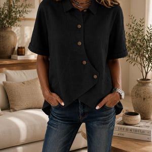 May include: A black short-sleeved shirt with a collar and a unique asymmetrical button-down front closure featuring light brown buttons. The shirt has a straight hemline and is paired with blue denim jeans, creating a casual and functional outfit.