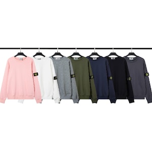 Può includere: Una fila di felpe girocollo in vari colori, tra cui rosa, bianco, grigio, verde oliva, blu navy, nero e antracite. Ogni felpa ha una piccola toppa sulla manica sinistra. Le felpe sono appese a un appendiabiti nero.