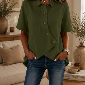 May include: An olive green short-sleeved shirt featuring a pointed collar, a button-down front with an asymmetrical overlapping hemline, and cuffed sleeves, paired with blue denim jeans and a silver-toned wristwatch on the wrist.