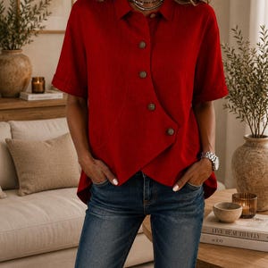 May include: A short-sleeved red button-down shirt featuring a unique asymmetrical front hemline and a classic collar. The shirt is crafted with a textured fabric and includes four visible buttons running down the front, paired with dark blue denim jeans.