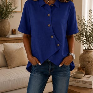 May include: A royal blue short-sleeved button-down shirt with a unique asymmetrical front hemline and a classic collar. The shirt features a row of brown buttons that run diagonally across the front, paired with dark blue denim jeans.