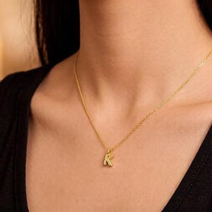 Puede incluir: Un collar dorado con un colgante con la forma de la letra "K". El collar lo lleva una persona con una camiseta sin mangas negra. El fondo está borroso, con tonos cálidos.