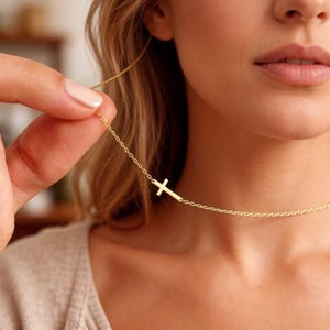 Puede incluir: Un delicado collar dorado con un colgante de cruz lateral. El collar tiene un diseño de cadena simple, y la cruz está colocada horizontalmente. La imagen es un primer plano, que destaca los finos detalles de la joya y los tonos cálidos del oro.