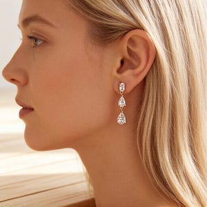 Puede incluir: Pendientes colgantes dorados con tres piedras de circonita cúbica transparente. Los pendientes tienen un diseño clásico con una pequeña piedra rectangular en la parte superior, seguida de dos piedras en forma de pera.