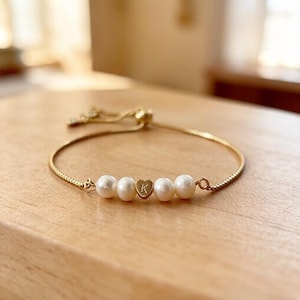 Puede incluir: Pulsera dorada con cuatro perlas blancas y un dije en forma de corazón con la letra "K". La pulsera ajustable está sobre una superficie de madera. Una joya delicada.