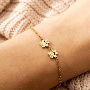 Puede incluir: Una pulsera dorada con dos colgantes de huellas de pata. Cada colgante tiene un nombre grabado. La pulsera está en una muñeca, con un suéter de punto beige en el fondo.