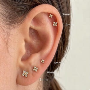 Puede incluir: Primer plano de una oreja adornada con múltiples pendientes de botón en forma de flor. Los pendientes son dorados con piedras claras y brillantes. Tamaños: 3 mm, 4 mm y 5 mm. La oreja está sobre un fondo neutro.