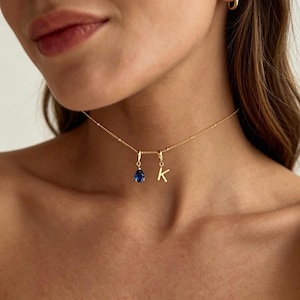 Puede incluir: Un collar dorado con una cadena delicada y dos colgantes. Uno es una piedra preciosa azul oscuro en forma de lágrima, el otro es la letra "K". El collar se lleva alrededor del cuello.