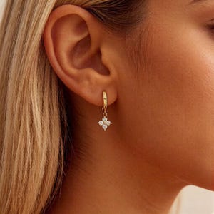 Puede incluir: Primer plano de un pendiente de aro dorado con un dije de flor de cuatro pétalos. El dije está adornado con pequeñas piedras preciosas transparentes. El pendiente se lleva en la oreja de una persona, con cabello rubio visible en el fondo.