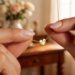 Puede incluir: Un collar dorado con un colgante en forma de corazón con la fecha grabada "4.3.24". El collar está sostenido entre dos manos. El fondo incluye un jarrón con flores y una mesa de madera.
