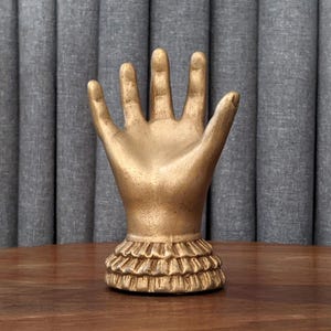 A small, decorative hand sculpture with a vintage gold finish. The hand is upright with an open palm and fingers spread, featuring a tiered, ruffled cuff at the wrist. It sits on a dark wooden tabletop against a textured grey pleated background.