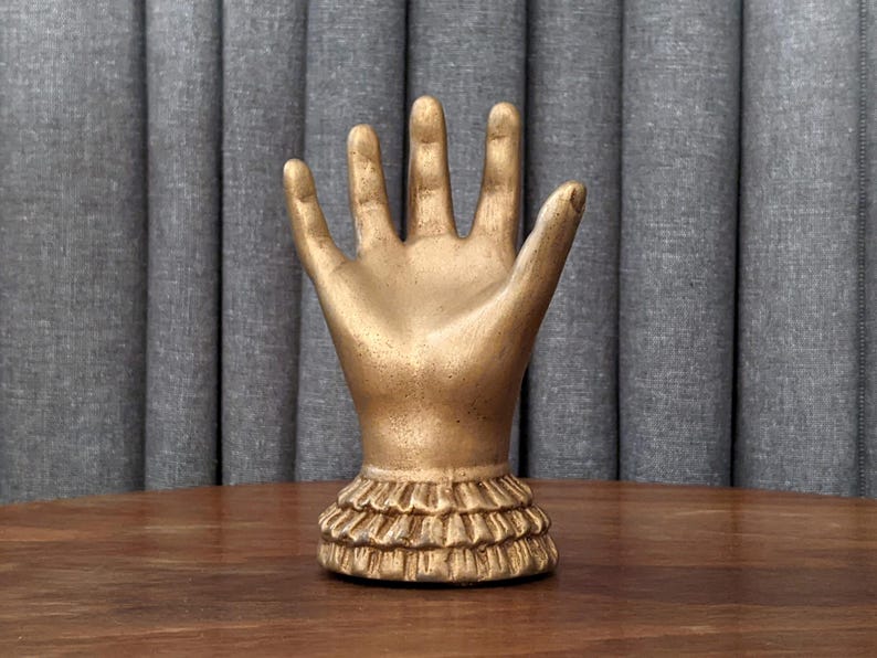 A small, decorative hand sculpture with a vintage gold finish. The hand is upright with an open palm and fingers spread, featuring a tiered, ruffled cuff at the wrist. It sits on a dark wooden tabletop against a textured grey pleated background.