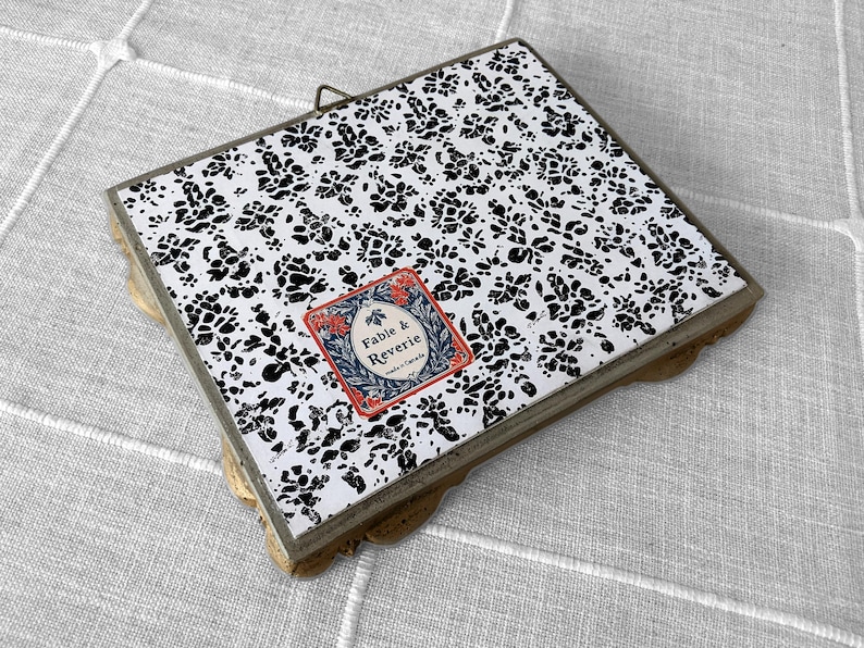 Op de afbeelding: Vierkant decoratief dienblad met een zwart-wit bloemenpatroon. De lade heeft een goudkleurige geschulpte rand en een klein label met de tekst "Fable & Reverie". Aan de bovenkant is een kleine metalen lus bevestigd om op te hangen.
